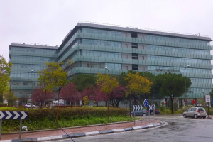 Großes modernes Gebäude mit zahlreichen Fenstern, als Hauptsitz der Europäischen Zentralbank identifiziert, umgeben von Grünflächen, Straßeninfrastruktur und Fahrzeugen unter einem bewölkten Himmel.