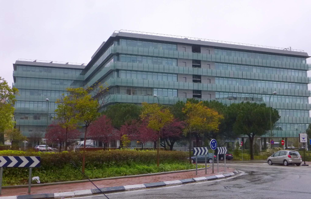 Großes modernes Gebäude mit zahlreichen Fenstern, als Hauptsitz der Europäischen Zentralbank identifiziert, umgeben von Grünflächen, Straßeninfrastruktur und Fahrzeugen unter einem bewölkten Himmel.