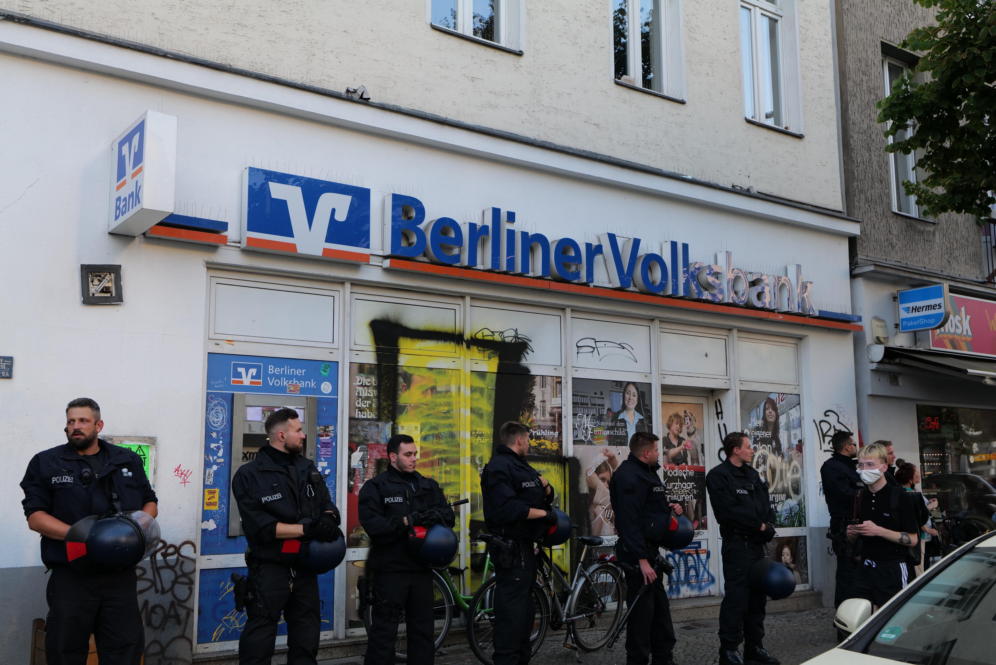 Eine Gruppe von Polizeibeamten vor einer Berliner Volksbank, einige mit Helmen, mit einem Auto auf der rechten Seite und einem Baum auf der linken Seite; Gebäude und Texttafeln im Hintergrund.