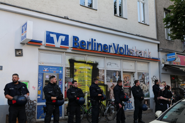 Eine Gruppe von Polizeibeamten vor einer Berliner Volksbank, einige mit Helmen, mit einem Auto auf der rechten Seite und einem Baum auf der linken Seite; Gebäude und Texttafeln im Hintergrund.