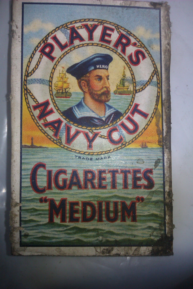 Ein Plakat an einer Wand mit einer Person in einer marineblauen Uniform mit weißem Hemd und roter Krawatte, die Arme verschränkt, strenger Gesichtsausdruck, mit Text, der "Player's Navy Cut Cigarettes Medium" in fetter schwarzer Schrift umgeben von einem weißen Rand liest.