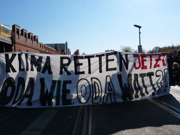 Eine Gruppe von Menschen steht auf der Straße und hält ein Schild hoch, auf dem steht "Klima Retten Jetz Oda Wie Oda Watt?" mit Gebäuden, Bäumen und einem Laternenpfahl im Hintergrund.
