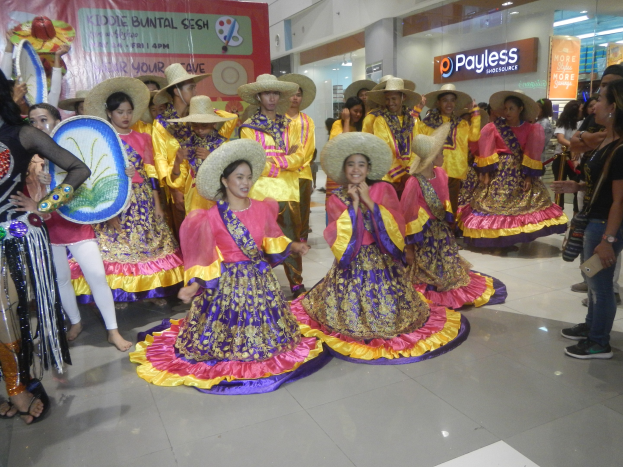 Eine Gruppe von Menschen in bunten Kleidern und Hüten, die in einem Einkaufszentrum tanzen, während andere Musikinstrumente halten und ein Banner trägt, bei einer likely feierlichen Szene, die Cinco de Mayo feiert.