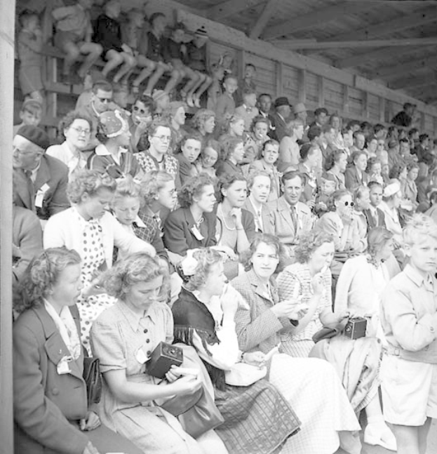 Schwarzes und weißes Foto einer Menge, die in einem Stadion sitzt und ein Sportereignis beobachtet, wobei einige Kameras halten und eine hölzerne Wand und ein Dach im Hintergrund zu sehen sind.