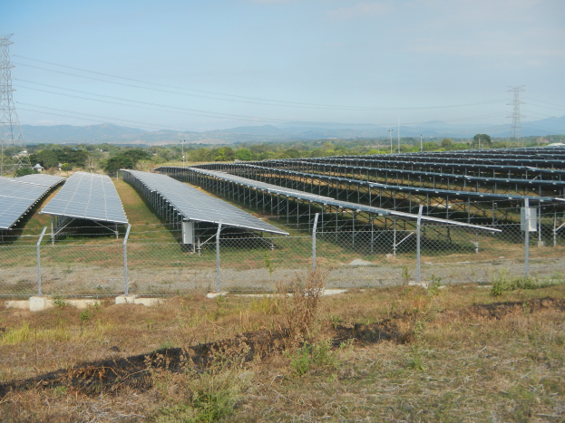 Ein Solarpanelfeld mit einem Zaun im Vordergrund, umgeben von Gras und Pflanzen, mit Bäumen, Übertragungsmasten mit Drähten und Hügeln im Hintergrund unter einem klaren blauen Himmel.