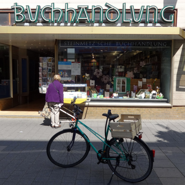 Eine Frau steht neben einem Fahrrad, das vor einem Geschäft geparkt ist, mit einem Fahrradrad im Vordergrund und durch die Glasscheibe des Geschäfts sichtbaren Büchern.