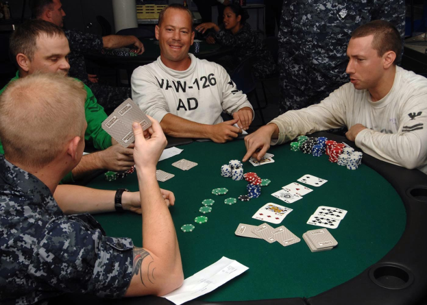 Eine Gruppe von Männern, die Poker an einem Tisch mit Münzen und Karten spielen, in einem Turnier-Setting mit einer Säule und anderen Gegenständen im Hintergrund.