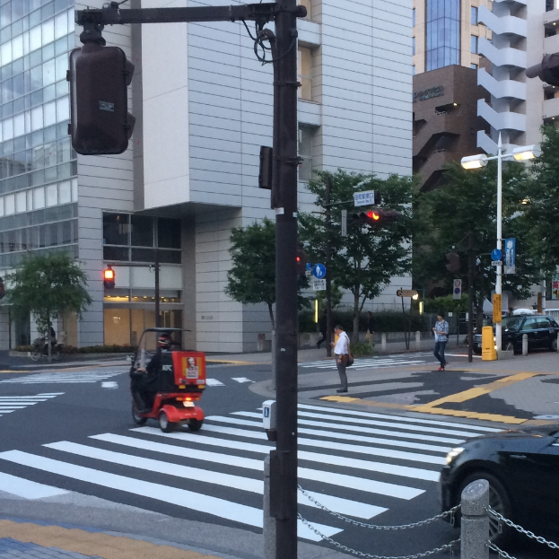 Ein Scooter auf der Straße mit einigen Personen im Hintergrund, eine Ampel im Vordergrund und Gebäude mit Bäumen in der Ferne.