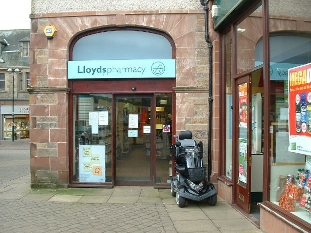 Apotheke mit einem Fahrzeug davor und einem Gebäude links daneben.