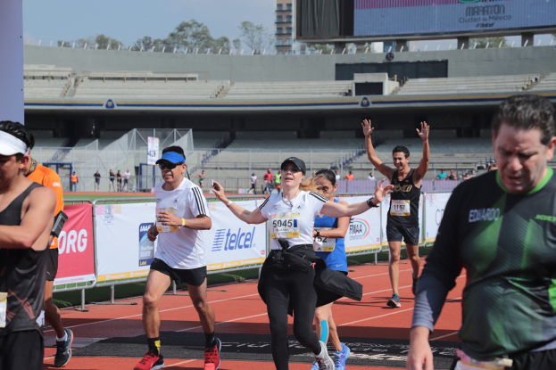 Gruppe von Läufern auf einer Außensportanlage mit Bannern, Anzeigetafel, Gebäude, Bäumen und bewölktem Himmel; einige Läufer tragen Turnschuhe und Hüte.