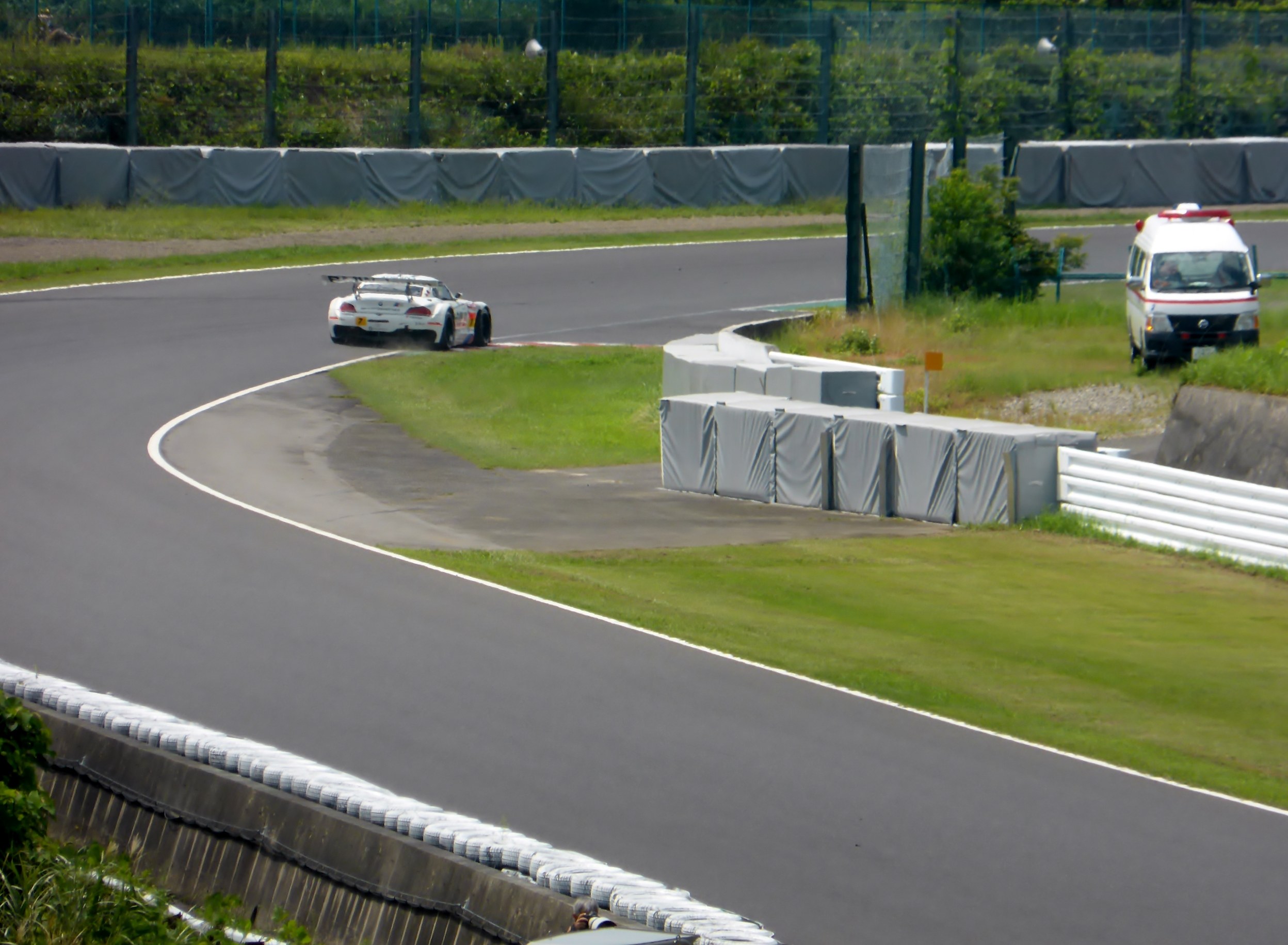 Ein Rennwagen fährt auf einer Rennstrecke mit einem Polizeiwagen im Hintergrund, umgeben von Gras, Pflanzen, Bäumen und Absperrungen, mit einem Fahrzeug auf der rechten Seite und Text unten.