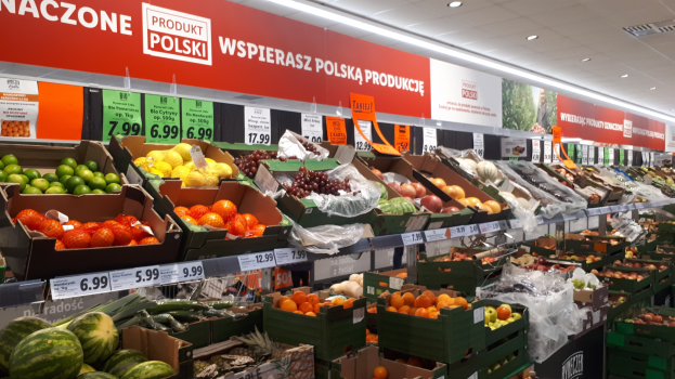 Ladeninneres mit verschiedenen Obst- und Gemüsesorten in Kartons mit Preisschildern, beleuchtet von Deckenlampen und mit informativen Tafeln an den Wänden.