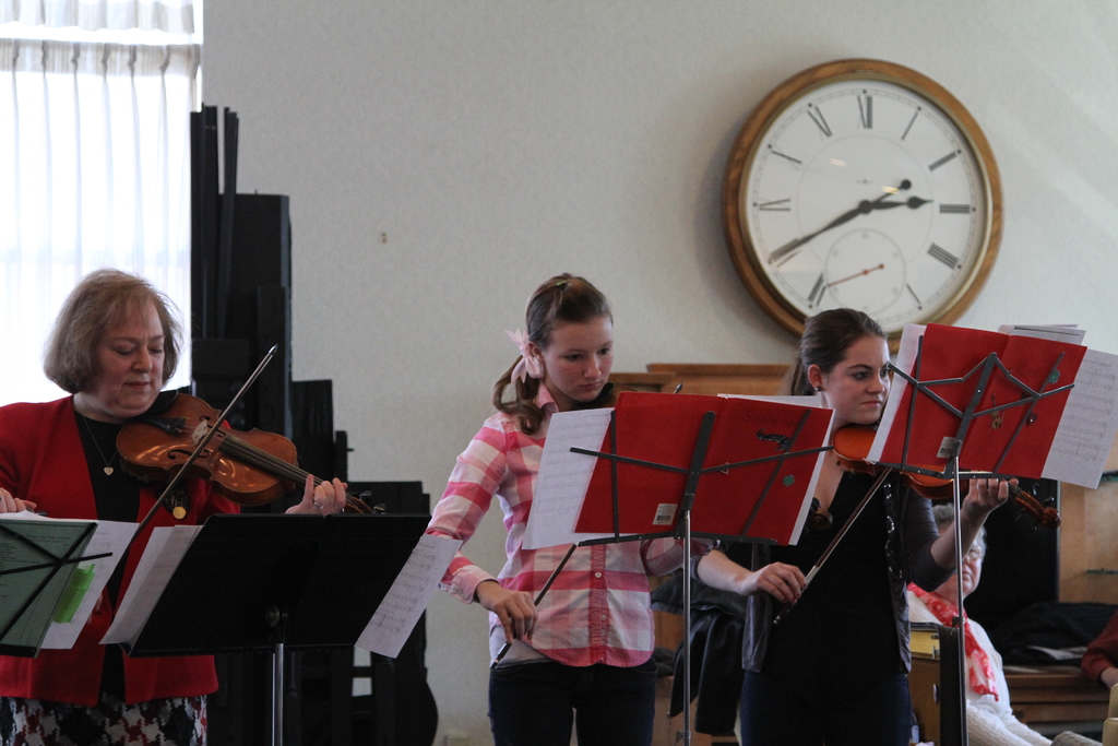 Drei Frauen, die auf der Bühne Geigen spielen, während eine Gruppe sitzender Frauen im Vordergrund zuhört; eine Uhr hängt an der weißen Wand im Hintergrund, mit einem weißen Vorhang in der linken Ecke.