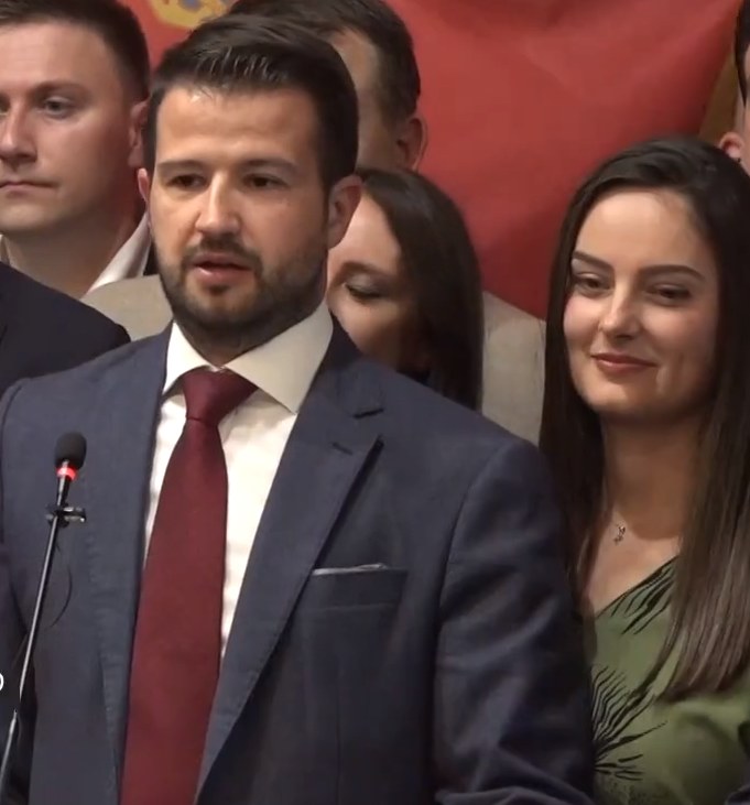 Ein Mann im Anzug spricht in ein Mikrofon an einem Rednerpult, mit einem Banner und einer Menge im Hintergrund, wahrscheinlich wegen eines Social-Media-Verbots.