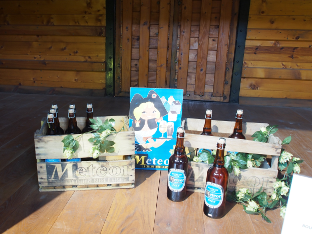 Ein hölzerner Kasten voller Bierflaschen auf einem hölzernen Boden in einer Gartenumgebung, mit Blättern und Blumen drumherum und einer hölzernen Wand und Tür im Hintergrund.