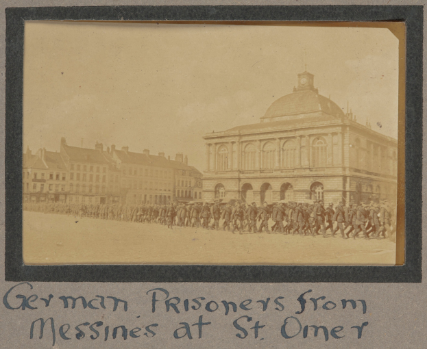 Eine Gruppe deutscher Gefangener aus Messines steht vor einem Geb"ude unter einem klaren blauen Himmel, mit Text am unteren Bildrand.