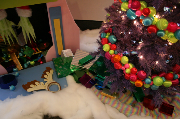 Drinnen, ein Weihnachtsbaum mit Lichterkette und Schmuck steht rechts, umgeben von Geschenkboxen und Dekorationsgegenständen, mit einer Hütte und Baumwollstoff oben links und Wolle unten.