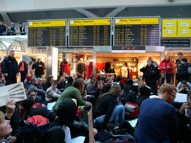 Eine große Gruppe von Menschen an einem Flughafen, einige sitzen mit Taschen und Papieren, andere stehen, mit Texttafeln, Schaufensterpuppen in Kleidern und Deckenleuchten im Hintergrund, was auf eine Demonstration hinweist.