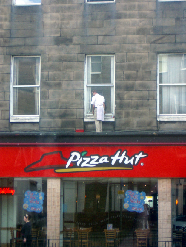 Ein Pizza Hut Shop mit einem Mann auf dem Dach, der die Glasscheiben reinigt und ein rotes Schild davor.