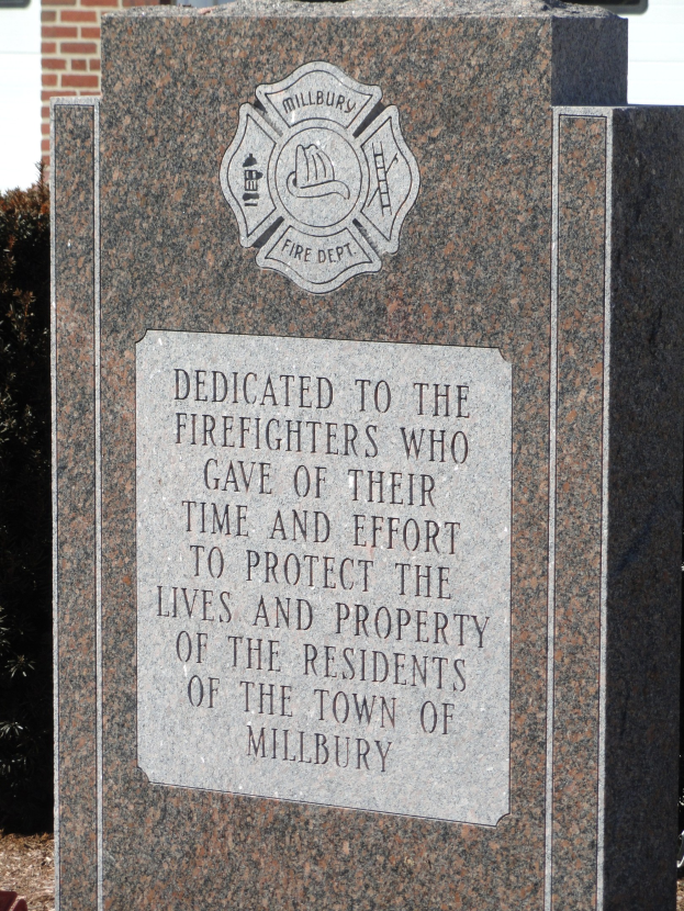 Denkmalstein mit einer Gedenktafel für Feuerwehrleute in Millbury, vor einem Backsteingebäude mit umgebender Vegetation.