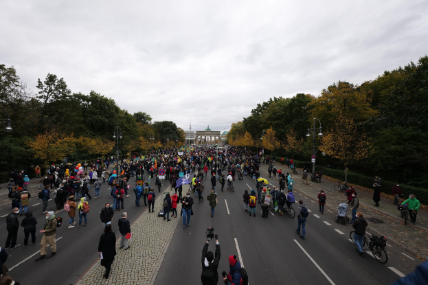 Eine große Menge marschiert auf einer baumbestandenen Straße in Berlin, hält Kameras während einer Protestaktion, mit einem Gebäude und einem klaren Himmel im Hintergrund.
