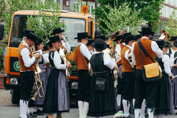 Eine Gruppe von Menschen in traditioneller bayrischer Tracht, die eine Straße entlangmarschieren, Instrumente spielen und Taschen tragen.