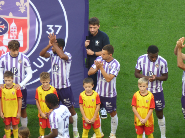 Eine Gruppe junger Jungs steht auf einem Fußballfeld, klatscht in die Hände, mit einem Banner und Logos im Hintergrund und saftigem grünem Gras darunter.
