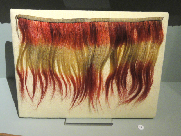 Nahaufnahme eines leuchtenden, bunt gef├Ąrbten Haarteil auf einem Brett in einer Museumsausstellung.