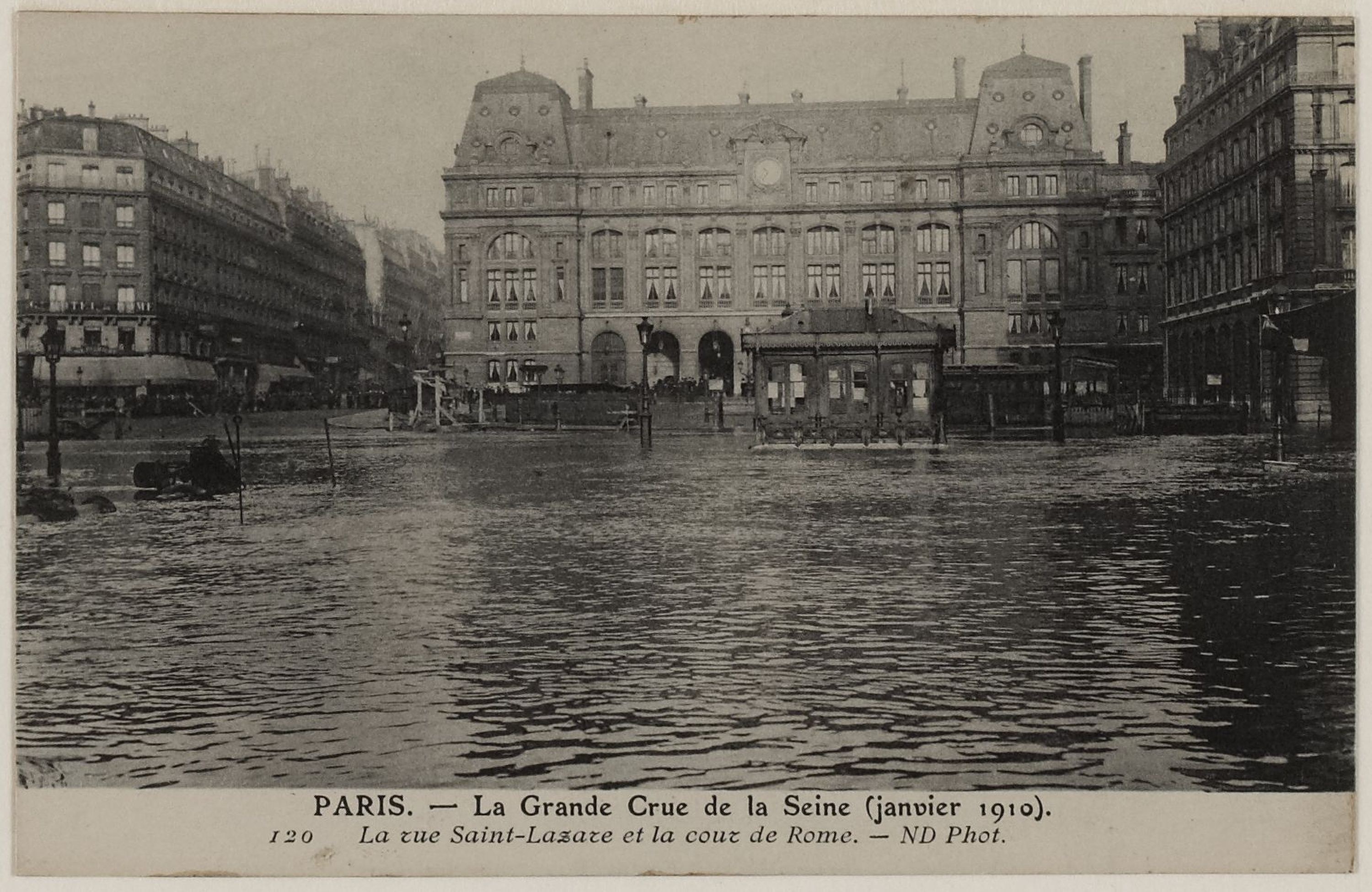 Schwarz-weiß-Foto einer überfluteten Straße in Paris mit Gebäuden, Straßenlaternen, Straßenlaternen, Kraftfahrzeugen und einem Himmel im Hintergrund, mit Text unten.