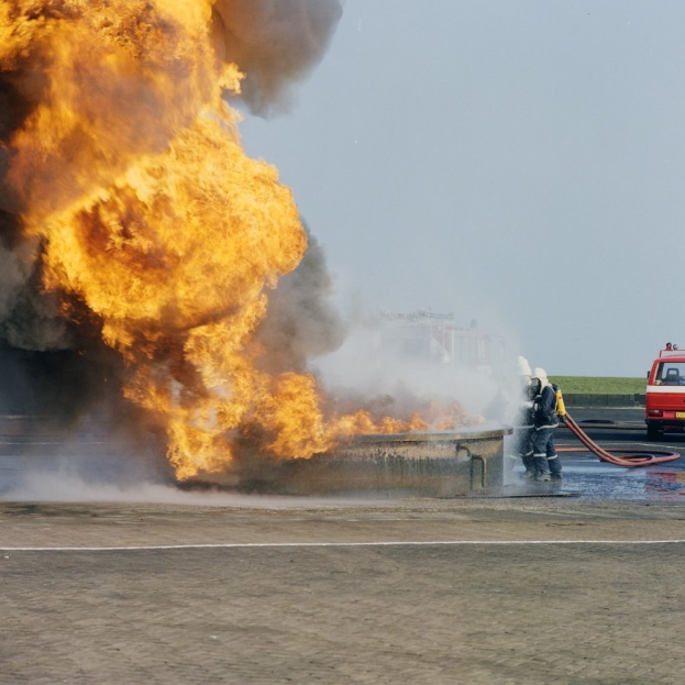 Löschfahrzeug von Flammen umgeben auf der Seite einer Straße mit zwei Helmträgern, die Rohre halten, einem Fahrzeug im Hintergrund und dem Himmel.
