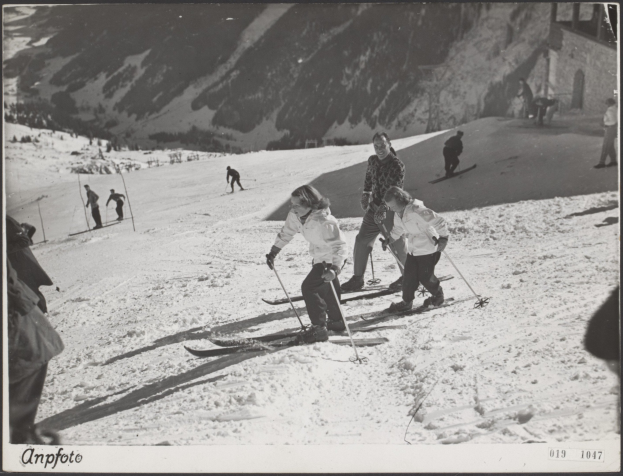 Gruppe von Menschen beim Skifahren auf einer schneebedeckten Piste mit Skistöcken, Hügeln und einem Gebäude im Hintergrund und Text am unteren Bildrand.