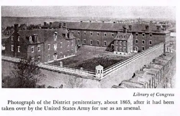 Ein Schwarz-Weiß-Foto des Bezirksgefängnisses aus dem Jahr 1865, das mehrere Gebäude, Bäume und eine Wand zeigt, mit Text am unteren Rand.