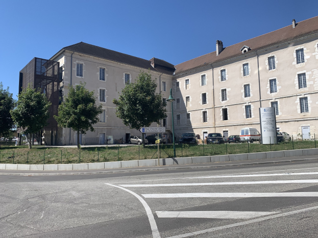 Großes Regierungsgebäude mit vielen Fenstern, umgeben von einem Zaun, Gras, Bäumen, Fahrzeugen, einer Straßenlaterne, einem Schild und einem bewölkten Himmel, identifiziert als Hauptquartier des französischen Gesundheits- und Sozialministeriums.