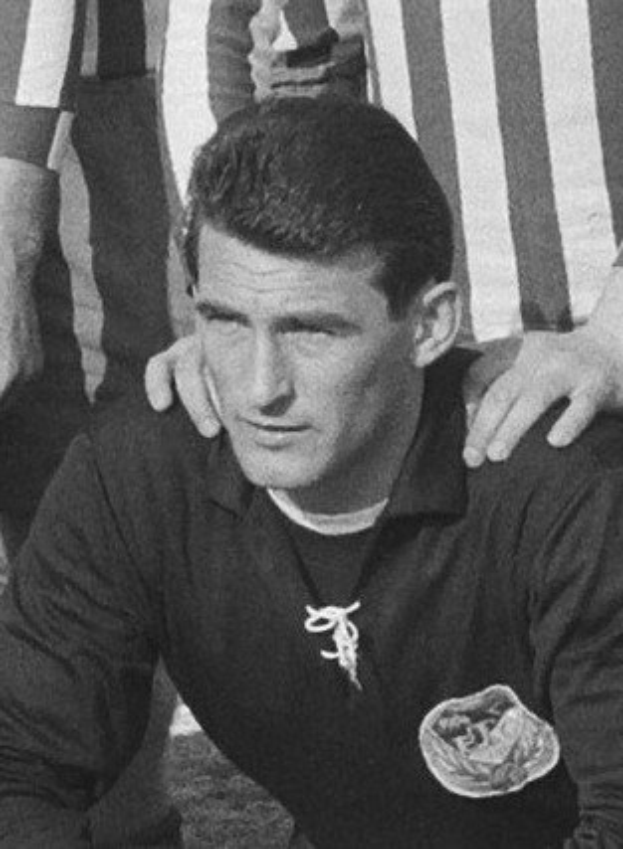Schwarzes und weißes Foto eines Mannes, der auf einem grünen Fussballfeld sitzend eine T-Shirt und eine Kette trägt, mit zwei anderen Personen, die hinter ihm stehen.