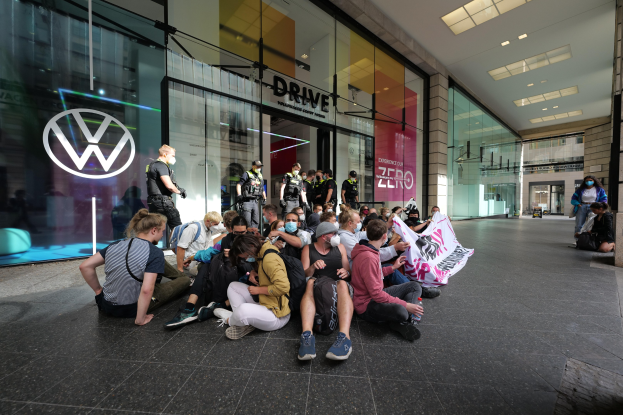 Eine Gruppe von Menschen sitzt vor einer Volkswagen-Vertretung mit Glaswänden auf dem Boden und hält eine Fahne mit Text, unter Deckenbeleuchtung, scheinbar protestierend.