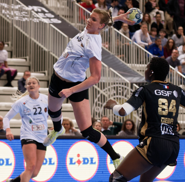 Eine Gruppe von Frauen, die Handball auf einem Platz spielt, mit einem Ball in der Luft, Zuschauern auf den Stufen und einem "Handball-Weltmeisterschaft 2019 - Tag 2"-Schild im Hintergrund.
