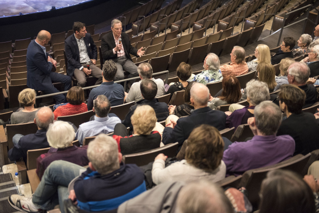 Gruppe von Menschen, die auf Stühlen sitzen und einer Bühne zugewandt sind, mit zwei Männern, die Mikrofone halten, und einer Treppe auf der linken Seite, die zur Bühne führt.