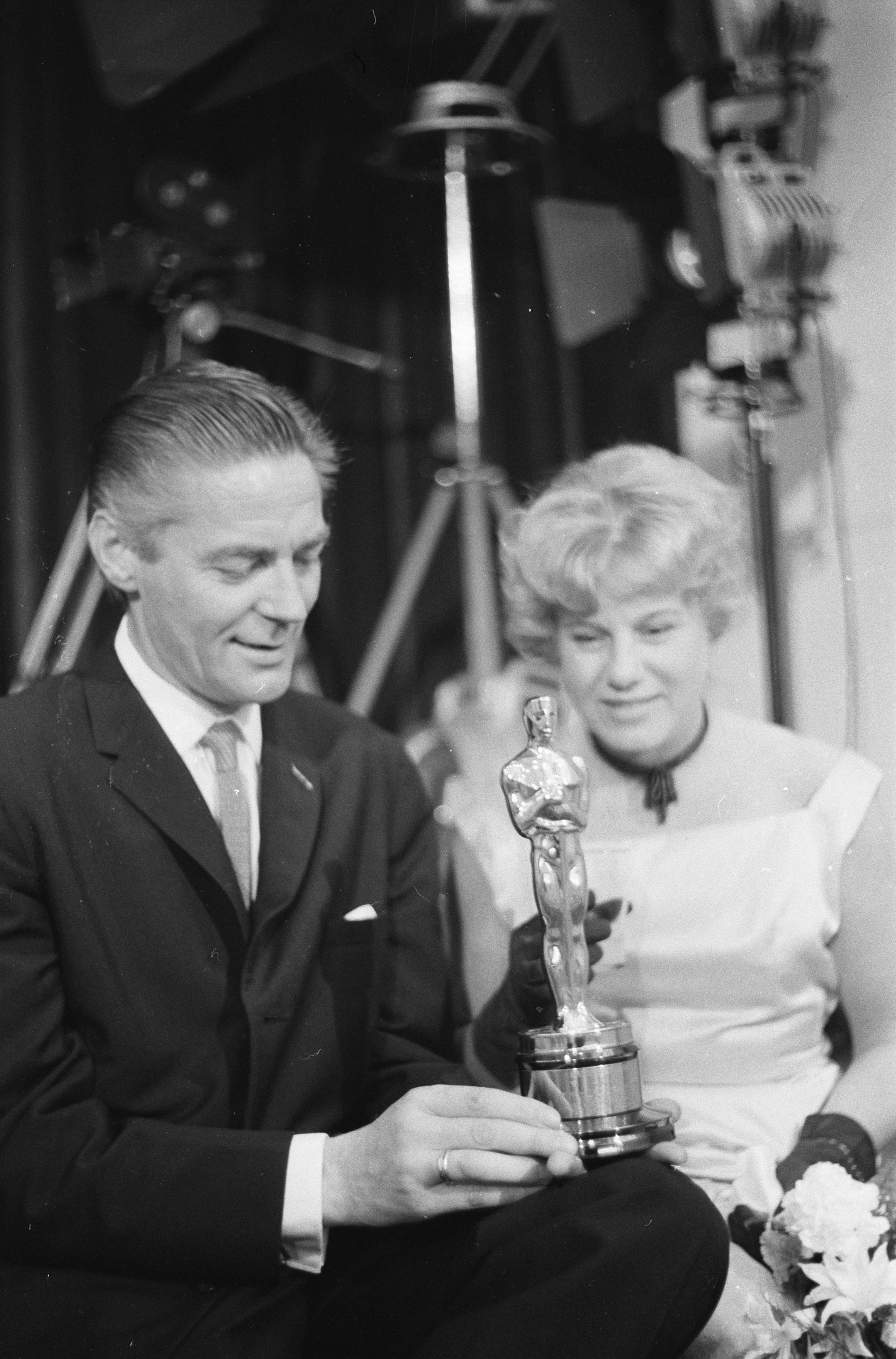 Ein Mann in einem schwarzen Blazer, weißem Hemd und Krawatte hält eine Oscar-Statue, während er neben einer Frau in Abendgarderobe sitzt, die einen Blumenstrauß hält, wobei Hintergrundlichter die schwarz-wei├če Szenerie beleuchten.