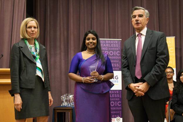 Zwei Frauen und ein Mann stehen auf einer Bühne, die Frau in der Mitte trägt ein purpurfarbenes Kleid und hält einen Award in der Hand, der Mann trägt ein schwarzes Sakko, ein weißes Hemd und eine rosafarbene Krawatte; ein brauner Vorhang ist dahinter, ein Podium mit einem Mikrofon links und mehrere Awards auf einem Tisch davor.
