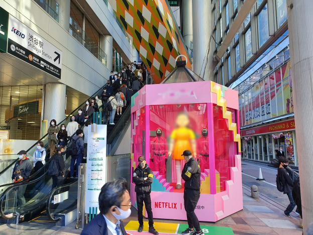Eine Gruppe von Menschen steht vor einem modernen Gebäude mit Glaswänden und Säulen, einige tragen Masken, mit einer Rolltreppe im Blickfeld. In der Mitte steht ein pinker Kasten mit Schaufensterpuppen und der Aufschrift "Netflix's new Netflix booth in Tokyo."