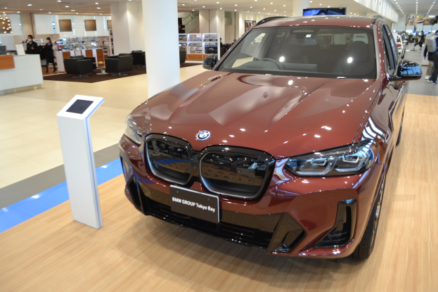 Roter BMW in hell erleuchteter Ausstellungshalle umgeben von Menschen, mit einem Tisch voller Gegenstände links und Sitzgelegenheiten im Hintergrund.