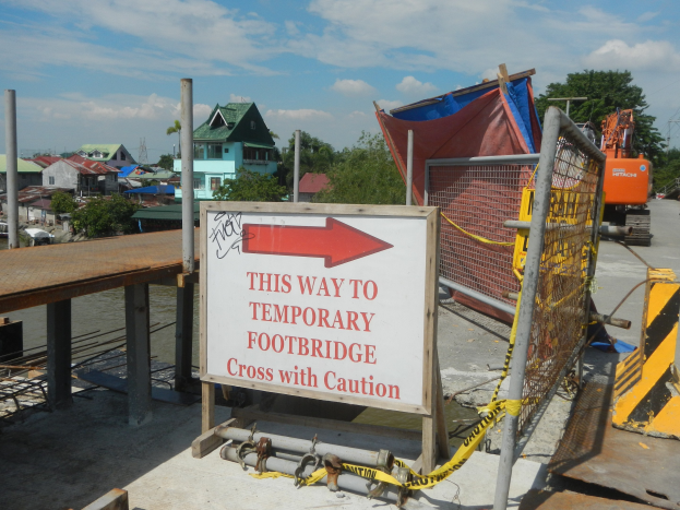 Temporäre Fußgängerbrücke mit Schild "Hier geht es zur temporären Fußgängerbrücke - Vorsicht beim Überqueren" mit Gebäuden, Bäumen und einem Fahrzeug im Hintergrund bei klarem blauem Himmel.