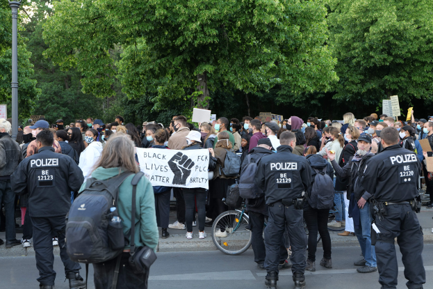 Eine große Gruppe von Menschen nimmt an einer Black Lives Matter Demonstration in Berlin teil, einige halten Schilder und andere tragen Kappen und Taschen, mit einem Fahrrad im Vordergrund und Bäumen und einem Pfahl im Hintergrund.