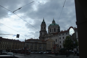 Gebäude in der Mitte, ein Mast und Bäume rechts, Autos, Menschen und ein Zug unten und Drähte oben.