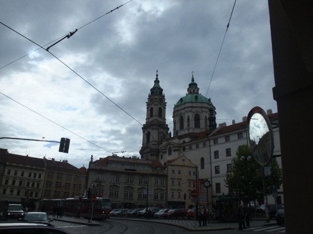 Gebäude in der Mitte, ein Mast und Bäume rechts, Autos, Menschen und ein Zug unten und Drähte oben.