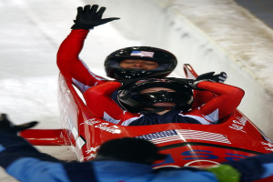 Zwei Bobsleighs fahren eine Bahn herunter, eines mit einem sichtbaren Helm und Handschuhen, mit einer Person unten und einer Wand im Hintergrund.