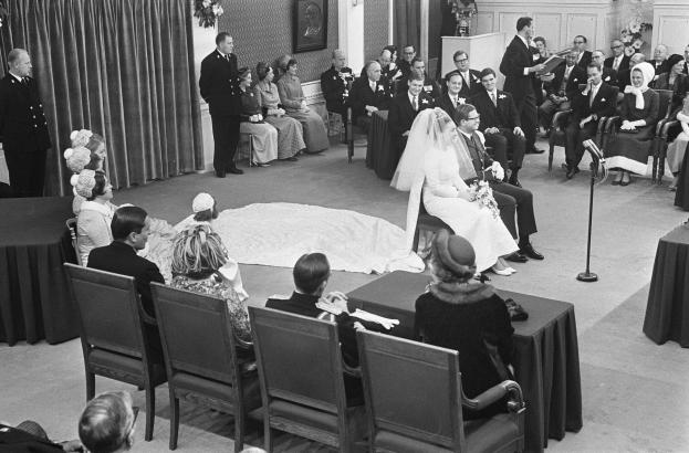 Schwarzes und weißes Foto einer Hochzeitszeremonie mit sitzenden und stehenden Menschen, einem Mann mit Mikrofon, einer Frau mit Blumenstrauß, einem Podium mit Mikrofon, Blumengestecken, einem Vorhang, einem Rahmen an der Wand und einer Tür im Hintergrund.