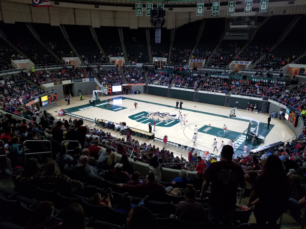 Eine große Arena voller Zuschauer, die ein Basketballspiel beobachten, mit einem Spielfeld in der Mitte und Anzeigetafeln, Lampen und anderen Gegenständen oben.