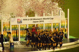 Eine Gruppe von Frauenfußballspielerinnen in schwarzen Uniformen feiert auf einer Bühne vor einem Publikum, mit einem dekorierten Hintergrund und einem grasbewachsenen Boden.