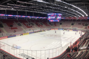 Ein Eishockeyspiel in einer großen Arena zwischen den St. Louis Blues und Detroit Red Wings, mit Zuschauern auf den Sitzplätzen und in der Nähe des Rinks, umgeben von einem Zaun, einem Display, Metallrahmen, Deckenleuchten und Dach.